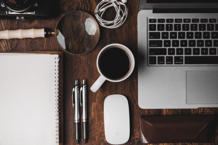 fotografia de mesa de trabalho com um computador, caderno, canetas, caneca de café, fones e uma lupa